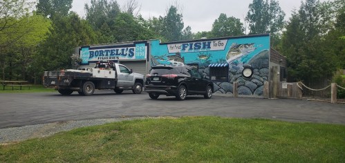 A fishery building with a mural, two parked vehicles, and a grassy area in front. Trees are visible in the background.