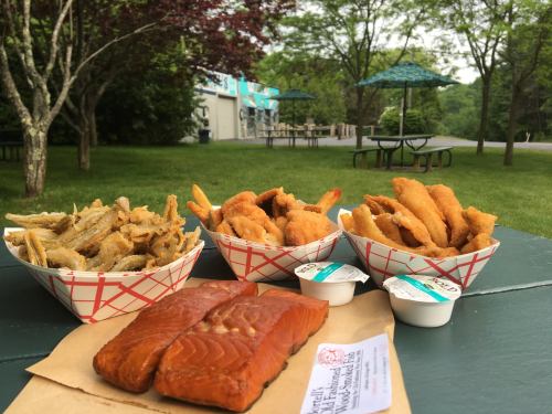 Two baskets of fried food and a portion of smoked salmon on a picnic table, surrounded by greenery.