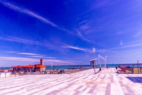 A wide, open waterfront plaza with a blue sky, featuring a red building and distant pier along the water.