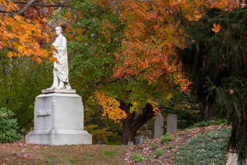 A white statue of a figure stands on a pedestal amidst vibrant autumn foliage and gravestones.