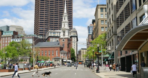 A busy city street with pedestrians, a dog, and historic buildings under a partly cloudy sky.
