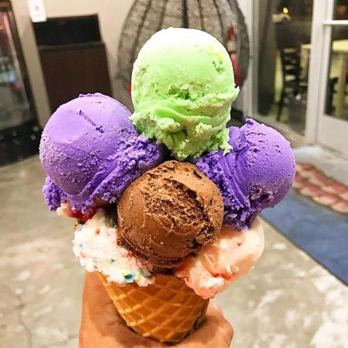 A hand holding a large ice cream cone with five colorful scoops: green, purple, brown, and two white with sprinkles.