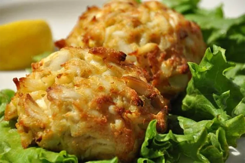 Two golden-brown crab cakes on a bed of fresh lettuce, accompanied by a lemon wedge.