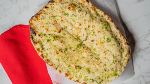 A large piece of garlic naan bread topped with herbs, resting on a white surface next to a red cloth.