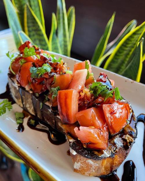 A slice of toasted bread topped with diced tomatoes, herbs, and balsamic glaze, served on a white plate with greenery.