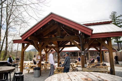 Two people converse under a wooden pavilion, surrounded by picnic tables and patrons enjoying the outdoor space.