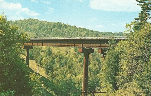 A highway bridge spans a lush green valley, supported by tall pillars, with mountains in the background under a blue sky.
