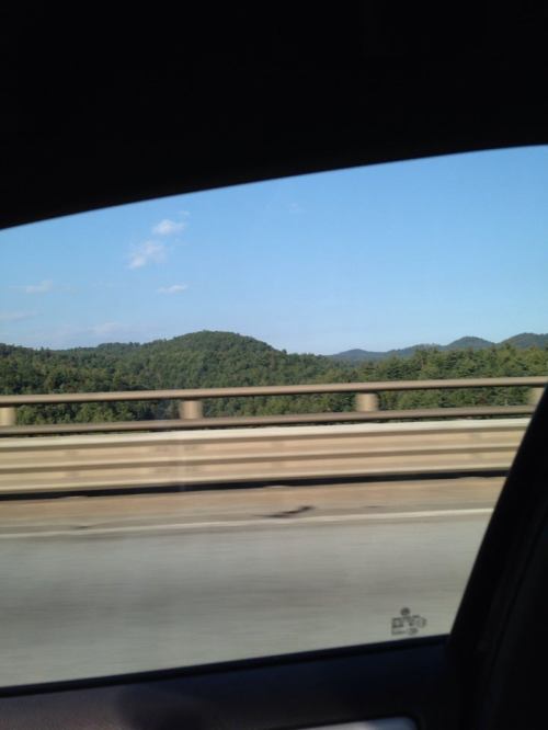 View of green mountains and blue sky from a car window, with a concrete barrier in the foreground.