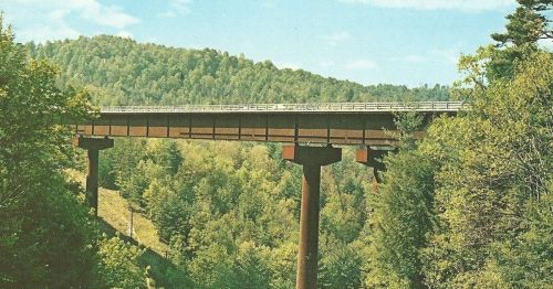 A highway bridge spans a lush green valley, surrounded by dense trees and rolling hills under a clear blue sky.