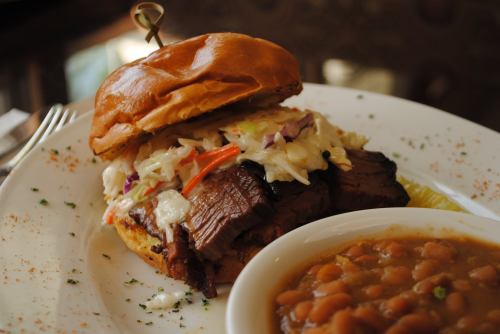 A close-up of a beef sandwich topped with coleslaw, served with a side of baked beans on a white plate.