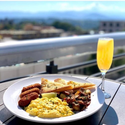 A plate of scrambled eggs, toast, and crispy potatoes, accompanied by a mimosa, with a scenic view in the background.