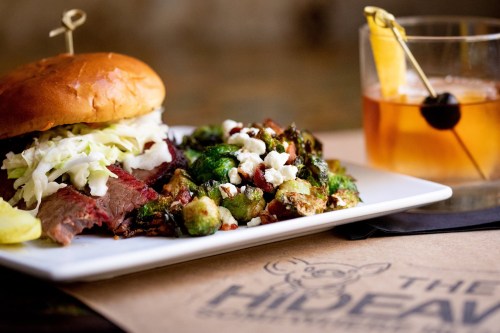A plate featuring a sandwich with coleslaw, a side of roasted vegetables, and a cocktail with a garnish.