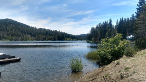 A serene lake surrounded by mountains and tall pine trees, with a small house visible in the distance.