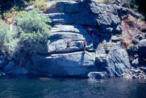 Two people on a rocky cliff by a river, one jumping into the water while another climbs the rocks.