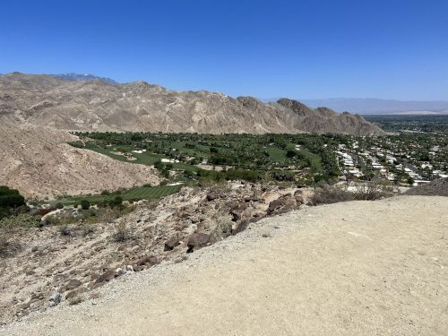 A panoramic view of a golf course nestled in a valley, surrounded by mountains under a clear blue sky.