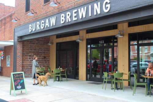 A woman walks a dog outside Burgaw Brewing, featuring outdoor seating and a chalkboard sign for soup and beer.