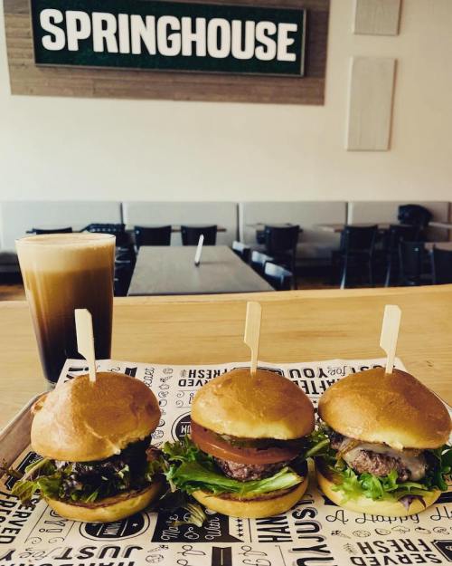 Three gourmet burgers on a tray with lettuce, tomato, and pickles, alongside a glass of iced coffee in a casual dining setting.