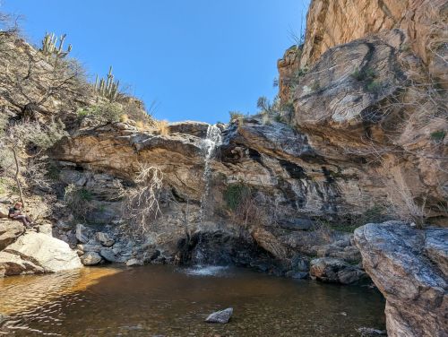 A serene waterfall cascading over rocks into a clear pool, surrounded by rugged cliffs and desert vegetation.