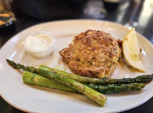 A plate featuring a crab cake, lemon wedge, and grilled asparagus with a side of dipping sauce.