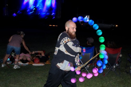 A person with a beard performs with colorful poi at a music festival, with others relaxing in the background.