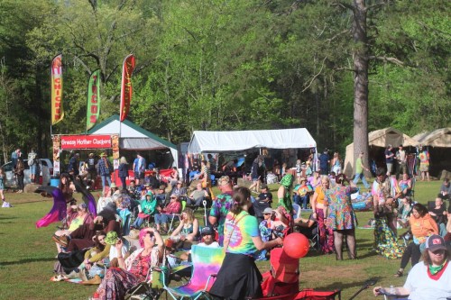 A vibrant outdoor festival scene with people in colorful attire, tents, and food stalls amidst green trees.