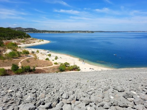 A scenic view of a beach by a calm lake, surrounded by greenery and hills under a clear blue sky.