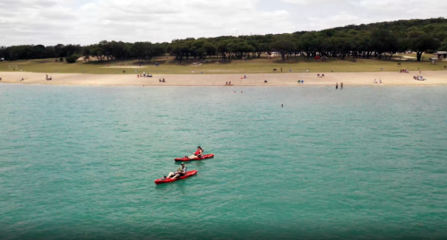Two people kayak on a turquoise lake, with a sandy beach and trees in the background under a partly cloudy sky.