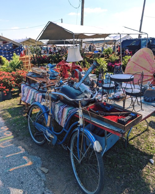 A blue vintage bicycle rests on a table filled with various antiques and decorative items at a bustling outdoor market.