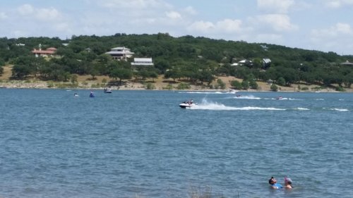 A sunny lake scene with people swimming and boats speeding across the water, surrounded by green hills and homes.