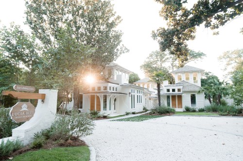 A charming coastal-style house with large windows, surrounded by trees and a gravel driveway, illuminated by sunlight.