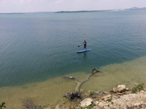 A person paddleboarding on calm water near a rocky shore with clear, shallow areas and distant hills.
