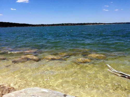 A serene lake scene with clear blue water, rocky shoreline, and a distant green landscape under a bright sky.