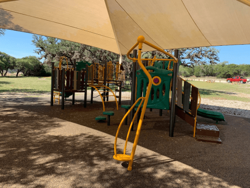 Playground equipment under a shaded structure, featuring climbing frames and slides, set in a grassy area.