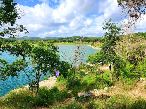 A scenic view of a lake surrounded by greenery and trees, with people enjoying the outdoors under a partly cloudy sky.