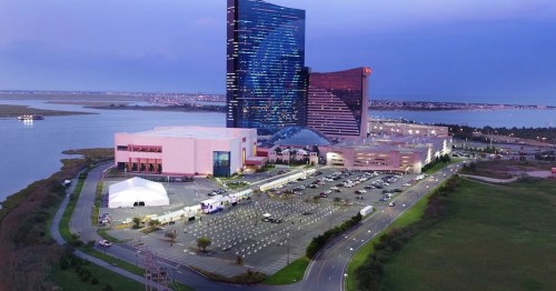 Aerial view of a modern casino complex by a waterfront, with a large parking lot and event tent in the foreground.