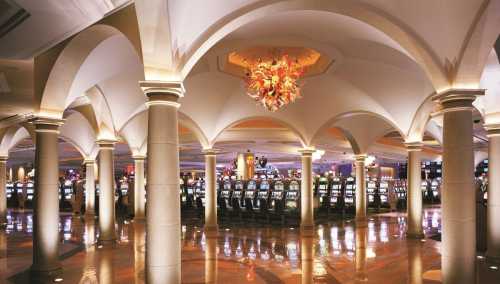 Elegant casino interior featuring arched columns, polished floors, and a chandelier, with rows of slot machines in the background.