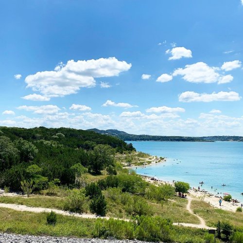 A scenic view of a beach by a lake, with people enjoying the water and lush greenery under a blue sky with clouds.