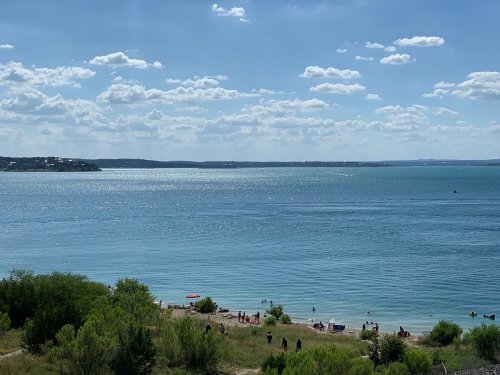 A serene lakeside view with people enjoying the beach, surrounded by greenery and a clear blue sky with fluffy clouds.