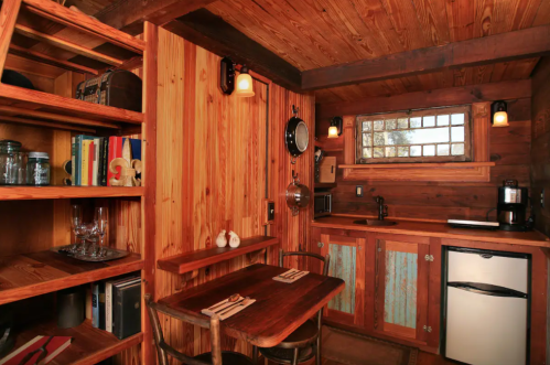 Cozy wooden cabin interior with a small dining table, kitchenette, shelves, and warm lighting.