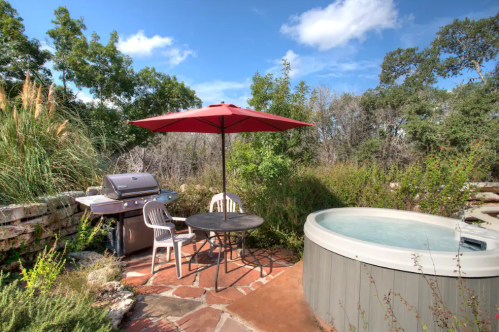 Outdoor patio with a grill, table, chairs, and a hot tub surrounded by greenery under a blue sky.