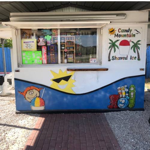Colorful shaved ice stand with a cheerful sun and cartoon characters painted on the exterior. Bright signage visible.
