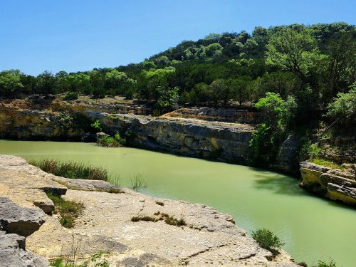 A serene river flows through rocky cliffs, surrounded by lush greenery under a clear blue sky.