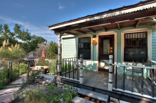 Charming light blue cottage with a porch, surrounded by greenery and a stone pathway, under a clear blue sky.