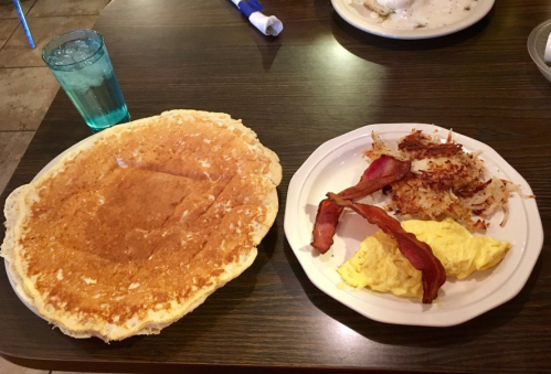 A large pancake next to a plate of scrambled eggs, bacon, and crispy hash browns, with a glass of water.