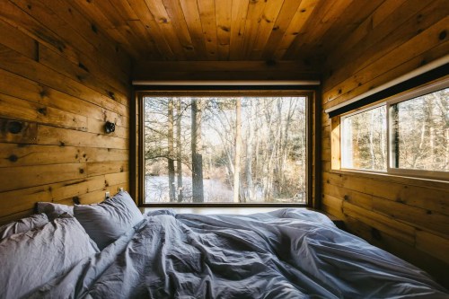 Cozy bedroom with wooden walls and large windows overlooking a serene forest landscape. Soft bedding is unmade.