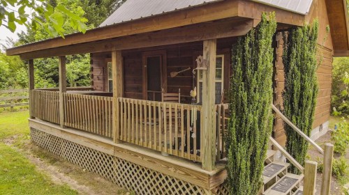 A rustic wooden cabin with a porch, surrounded by greenery and a wooden fence.