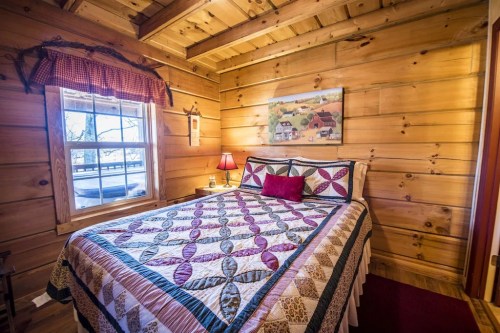 Cozy wooden bedroom with a quilted bedspread, a window, and rustic decor, featuring a painting on the wall.