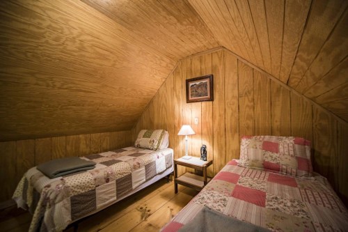 Cozy attic bedroom with two twin beds, wooden walls, a lamp, and a framed picture.