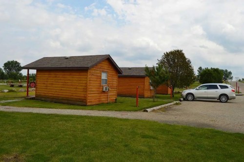 Two wooden cabins with a car parked nearby, set in a grassy area under a cloudy sky.