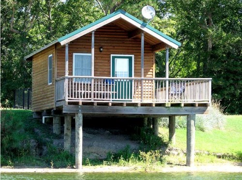 A wooden cabin on stilts by a lake, featuring a porch and green roof surrounded by trees.